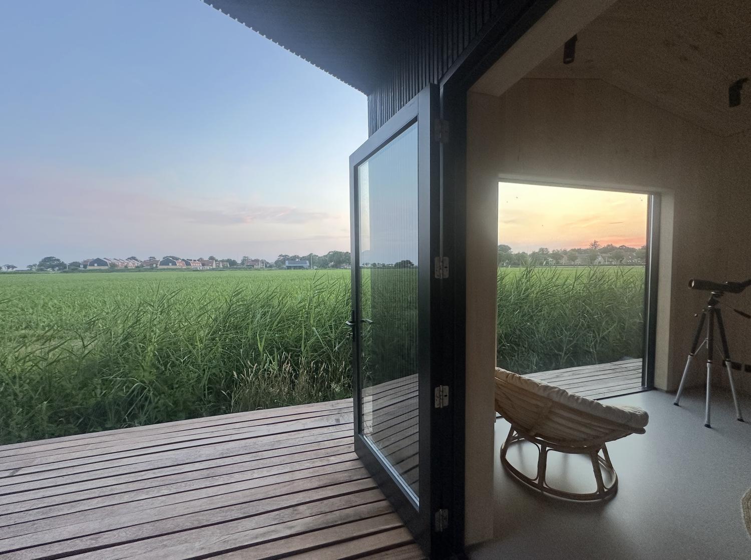 Openslaande zwarte deur naar de vlonder, met rieten fauteuil en telescoop in het lichte houten interieur