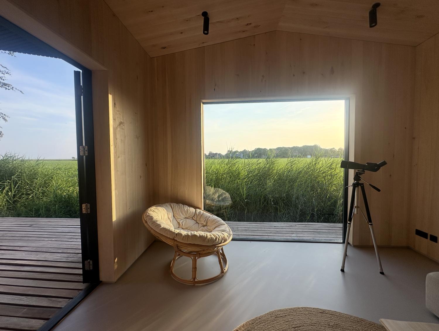 Rieten fauteuil voor een groot panoramaraam met uitzicht op het rietveld, naast een telescoop